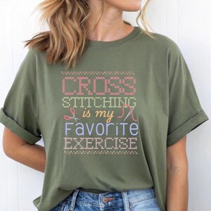 Op de afbeelding: Een groen T-shirt met de tekst "Cross Stitching is my Favorite Exercise" in een kruissteek-stijl lettertype. De tekst wordt omringd door een rand van kleine rode, gele en blauwe x'en.