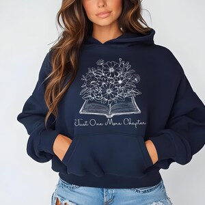 Op de afbeelding: Een marineblauwe hoodie met een witte afbeelding van een boek met bloemen erop en de tekst "Just One More Chapter".