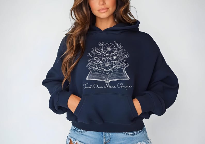 Op de afbeelding: Een marineblauwe hoodie met een witte afbeelding van een boek met bloemen erop en de tekst "Just One More Chapter".