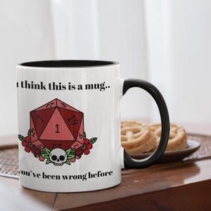 May include: White ceramic mug with a black interior and handle. The mug features a red, geometric die design with the number 1, surrounded by flowers and a skull. Text on the mug reads "I think this is a mug.." and "you've been wrong before."