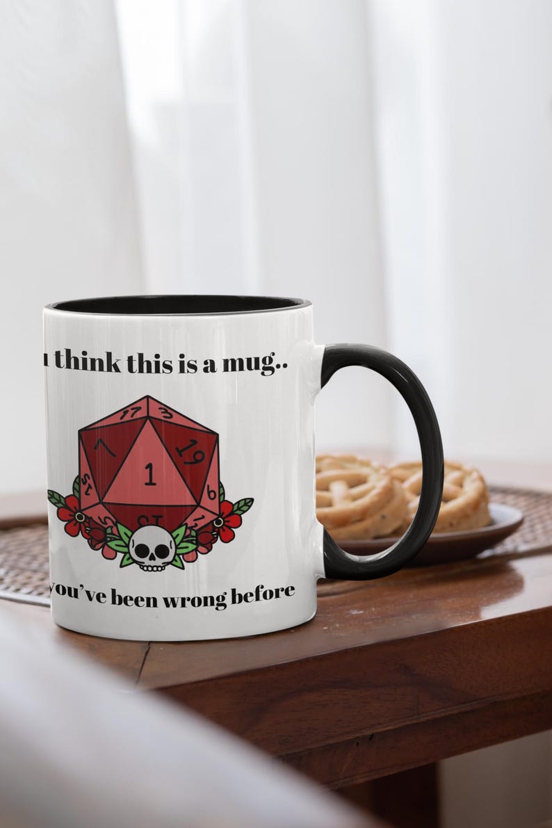 May include: White ceramic mug with a black interior and handle. The mug features a red, geometric die design with the number 1, surrounded by flowers and a skull. Text on the mug reads "I think this is a mug.." and "you've been wrong before."
