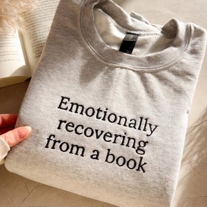 Może przedstawiać: Jasnoszara bluza z haftowanym napisem "Emotionally recovering from a book". Bluza z okrągłym dekoltem jest złożona. Książka i dekoracyjna roślina znajdują się w tle. Bluza leży na jasnej powierzchni.