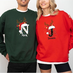 Op de afbeelding: Twee kersttruien, één groene en één rode. De groene trui heeft de witte letter "N" met een kerstmuts en de naam "Nick". De rode trui heeft de zwarte letter "J" met een kerstmuts en de naam "Jasmin".