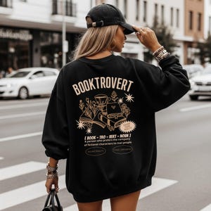 Puede incluir: Sudadera negra con la palabra "BOOKTROVERT" en color beige claro. La parte trasera de la sudadera presenta un gráfico de libros, plantas y estrellas. Debajo está la definición de "BOOKTROVERT".