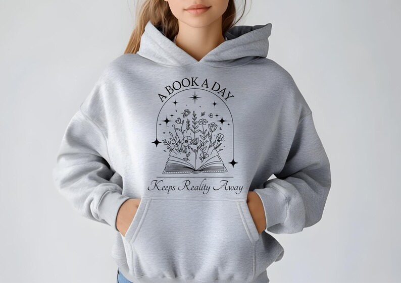 Puede incluir: Una sudadera con capucha gris con el texto "A Book A Day Keeps Reality Away" y una ilustraci&oacute;n en blanco y negro de un libro con flores y estrellas.