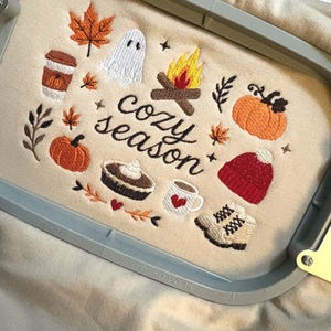 Geborduurde beige sweatshirt met het opschrift 'Cozy Season' en een herfstig borduurontwerp van een kampvuur, pompoenen, spookje, pumpkin spice koffie, warme chocolademelk, herfstbladeren, taart, gebreide muts en winterlaarsjes. De sweater ligt op een open boek met dennenappels, kaarslicht en herfstdecoratie. Perfecte herfsttrui voor Halloween, cozy season en herfstliefhebbers.