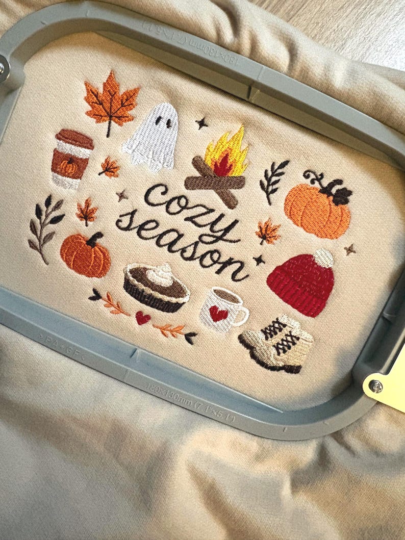 Geborduurde beige sweatshirt met het opschrift 'Cozy Season' en een herfstig borduurontwerp van een kampvuur, pompoenen, spookje, pumpkin spice koffie, warme chocolademelk, herfstbladeren, taart, gebreide muts en winterlaarsjes. De sweater ligt op een open boek met dennenappels, kaarslicht en herfstdecoratie. Perfecte herfsttrui voor Halloween, cozy season en herfstliefhebbers.