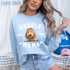Może przedstawiać: Jasnoniebieska bluza z napisem "DOODLE MAMA EST. 2024" i grafiką psa goldendoodle. Bluza ma odciski łap i jest wykonana z miękkiego materiału.