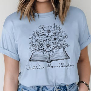 Op de afbeelding: Een lichtblauw t-shirt met een zwart-witte illustratie van een open boek met bloemen erop. De tekst "Just One More Chapter" is onder het boek gedrukt.