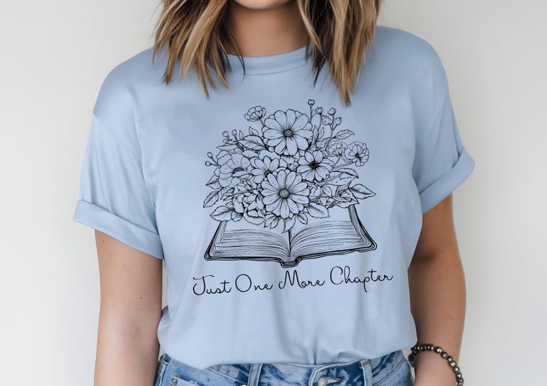 Op de afbeelding: Een lichtblauw t-shirt met een zwart-witte illustratie van een open boek met bloemen erop. De tekst "Just One More Chapter" is onder het boek gedrukt.