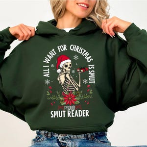 Peut inclure: Sweat à capuche vert foncé avec un motif festif. Il présente un squelette portant un bonnet de Père Noël tenant un cadeau, avec le texte "All I Want For Christmas Is Smut" et "Proud Smut Reader". Le motif comprend des flocons de neige et des poinsettias.