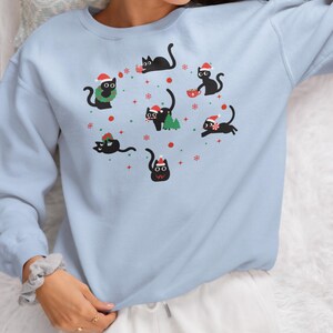 K&ouml;nnte beinhalten: Ein hellblauer Pullover mit einem festlichen Design, das schwarze Katzen zeigt, die Weihnachtsm&uuml;tzen tragen und weihnachtliche Gegenst&auml;nde halten. Die Katzen sind von Schneeflocken und anderen Weihnachtsdekorationen umgeben.