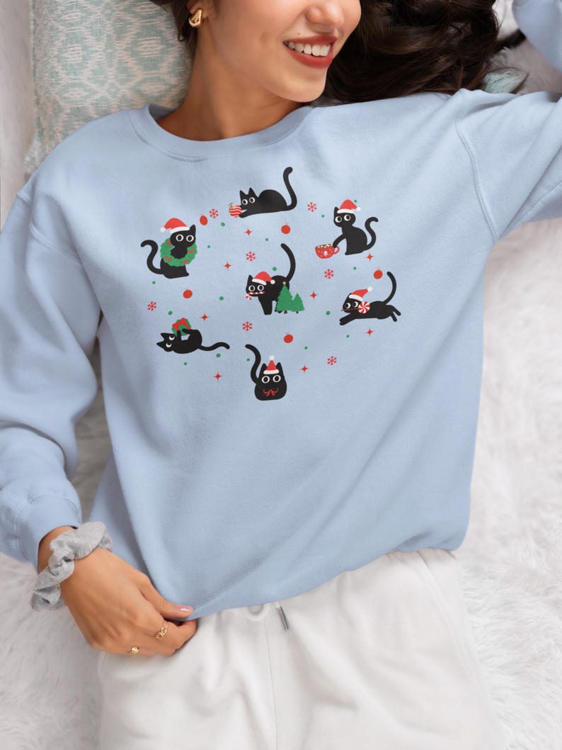 K&ouml;nnte beinhalten: Ein hellblauer Pullover mit einem festlichen Design, das schwarze Katzen zeigt, die Weihnachtsm&uuml;tzen tragen und weihnachtliche Gegenst&auml;nde halten. Die Katzen sind von Schneeflocken und anderen Weihnachtsdekorationen umgeben.