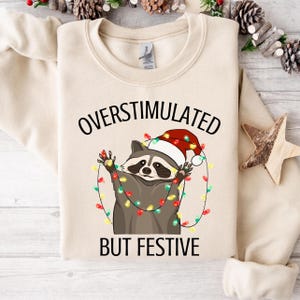 Puede incluir: Sudadera beige con el texto "OVERSTIMULATED BUT FESTIVE". El gráfico presenta un mapache con un gorro de Papá Noel y envuelto en luces navideñas. La sudadera tiene cuello redondo y mangas largas.