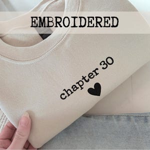 Op de afbeelding: Een beige sweatshirt met het woord "EMBROIDERED" bovenaan. Daaronder staan de woorden "chapter 30" gedrukt met een zwart hart. De sweatshirt is opgevouwen en ligt op een spijkerbroek.