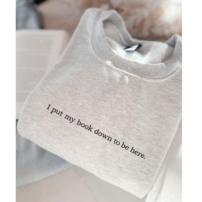May include: A light gray sweatshirt with the text "I put my book down to be here." The sweatshirt is folded and appears to be made of a soft material. The text is in a simple, black font.