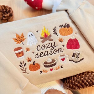 Geborduurde beige sweatshirt met het opschrift 'Cozy Season' en een herfstig borduurontwerp van een kampvuur, pompoenen, spookje, pumpkin spice koffie, warme chocolademelk, herfstbladeren, taart, gebreide muts en winterlaarsjes. De sweater ligt op een open boek met dennenappels, kaarslicht en herfstdecoratie. Perfecte herfsttrui voor Halloween, cozy season en herfstliefhebbers.