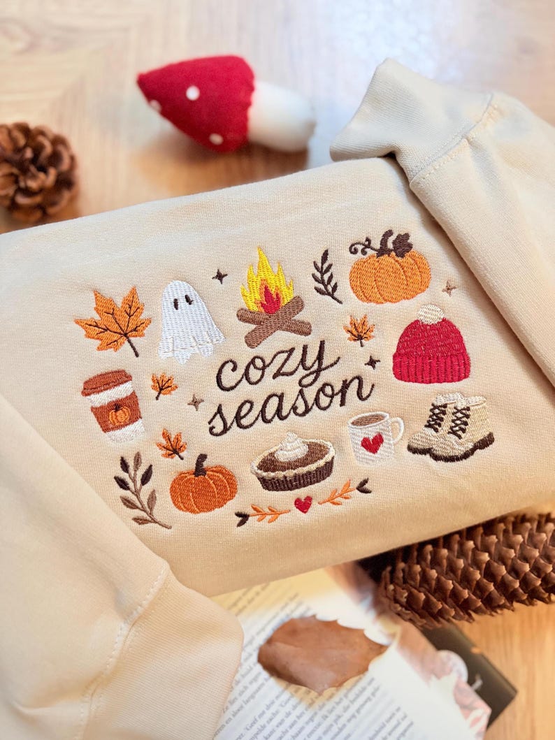Geborduurde beige sweatshirt met het opschrift 'Cozy Season' en een herfstig borduurontwerp van een kampvuur, pompoenen, spookje, pumpkin spice koffie, warme chocolademelk, herfstbladeren, taart, gebreide muts en winterlaarsjes. De sweater ligt op een open boek met dennenappels, kaarslicht en herfstdecoratie. Perfecte herfsttrui voor Halloween, cozy season en herfstliefhebbers.