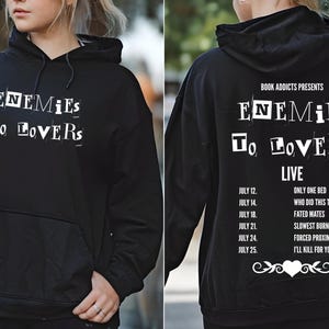 Peut inclure: Sweat-shirt à capuche noir avec du texte blanc qui dit "Enemies to Lovers Live" et une liste de dates et de titres de spectacles.