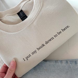 May include: A beige sweatshirt with the text "I put my book down to be here" in black. The sweatshirt is folded, with a portion of blue jeans visible. The Gildan tag is visible.