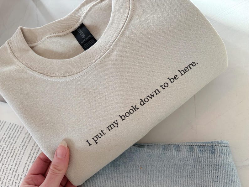 May include: A beige sweatshirt with the text "I put my book down to be here" in black. The sweatshirt is folded, with a portion of blue jeans visible. The Gildan tag is visible.