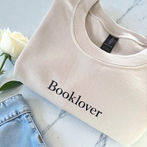 Può includere: Una felpa beige girocollo con la scritta "Booklover" stampata in nero. La felpa è piegata, con una rosa bianca e una porzione di jeans blu visibili sullo sfondo.