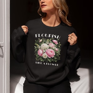 Op de afbeelding: Zwart sweatshirt met een bloemenontwerp met roze en witte pioenrozen en groene bladeren. De tekst "FLOURISH LIKE A FLOWER" is in het wit boven en onder het bloemstuk gedrukt.