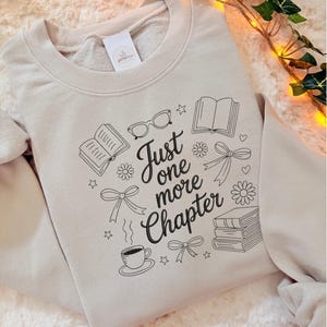Op de afbeelding: Een beige sweater met ronde hals voorzien van zwarte lijntekeningen van boeken, een bril, strikjes, bloemen, sterren en een koffiekopje. De tekst "Just one more Chapter" staat in het midden gedrukt in een zwart cursief lettertype.