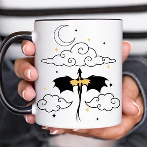 Op de afbeelding: Witte keramische mok met een zwarte handgreep en rand. De mok heeft een zwart draken silhouet met gouden accenten, vliegend tussen wolken, sterren en een halve maan. Het ontwerp staat tegen een witte achtergrond.