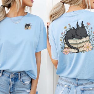 Op de afbeelding: Twee lichtblauwe T-shirts met een zwarte draak afbeelding. De draak slaapt op een stapel boeken met bloemen eromheen. Eén shirt heeft de afbeelding aan de voorkant en de andere aan de achterkant.