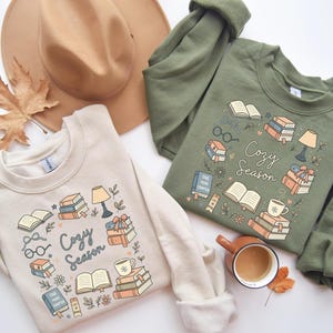 Op de afbeelding: Twee sweatshirts, één beige en één olijfgroen, met een gezellig leesthema. Elk sweatshirt heeft een ontwerp van boeken, brillen, een lamp en een koffiekopje, met de woorden "Cozy Season". Een bruine hoed en een kopje koffie staan ook op de foto.