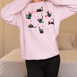 K&ouml;nnte beinhalten: Ein rosa Sweatshirt mit einem festlichen Design mit schwarzen Katzen, die Weihnachtsm&uuml;tzen tragen und weihnachtliche Gegenst&auml;nde halten. Die Katzen sind von Schneeflocken und anderen Weihnachtsdekorationen umgeben.
