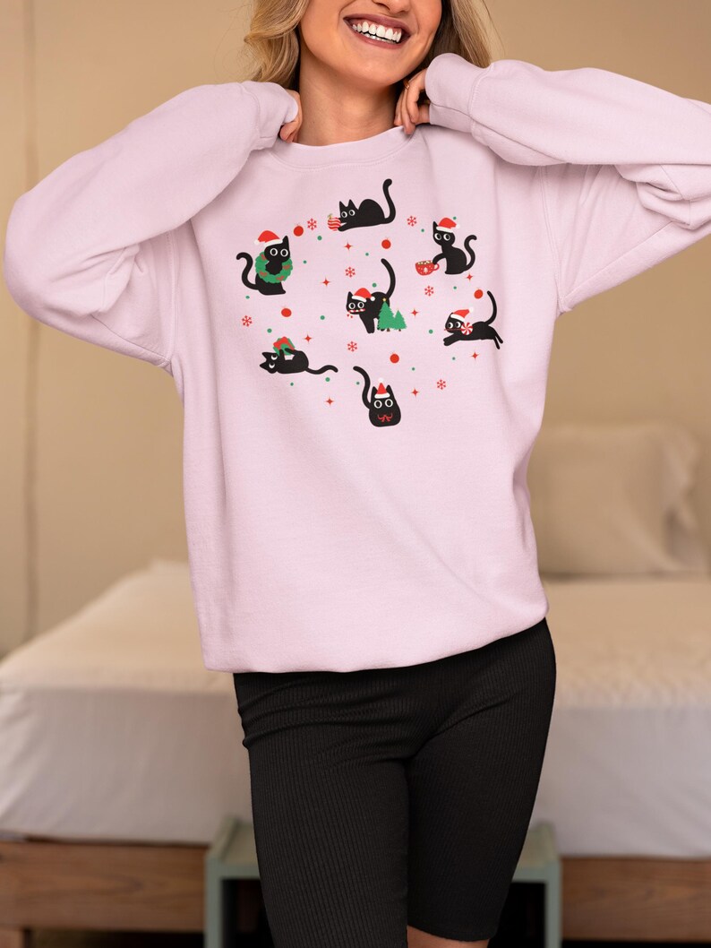 K&ouml;nnte beinhalten: Ein rosa Sweatshirt mit einem festlichen Design mit schwarzen Katzen, die Weihnachtsm&uuml;tzen tragen und weihnachtliche Gegenst&auml;nde halten. Die Katzen sind von Schneeflocken und anderen Weihnachtsdekorationen umgeben.