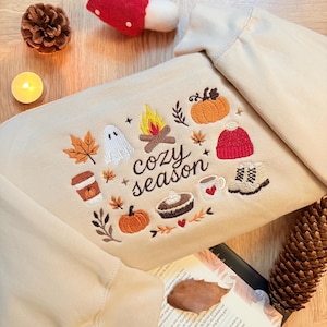 Geborduurde beige sweatshirt met het opschrift 'Cozy Season' en een herfstig borduurontwerp van een kampvuur, pompoenen, spookje, pumpkin spice koffie, warme chocolademelk, herfstbladeren, taart, gebreide muts en winterlaarsjes. De sweater ligt op een open boek met dennenappels, kaarslicht en herfstdecoratie. Perfecte herfsttrui voor Halloween, cozy season en herfstliefhebbers.