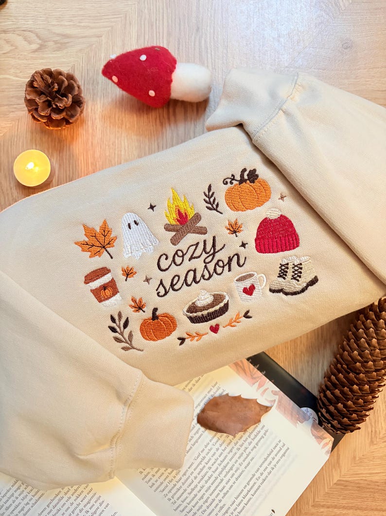 Geborduurde beige sweatshirt met het opschrift 'Cozy Season' en een herfstig borduurontwerp van een kampvuur, pompoenen, spookje, pumpkin spice koffie, warme chocolademelk, herfstbladeren, taart, gebreide muts en winterlaarsjes. De sweater ligt op een open boek met dennenappels, kaarslicht en herfstdecoratie. Perfecte herfsttrui voor Halloween, cozy season en herfstliefhebbers.
