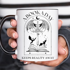 Op de afbeelding: Een witte keramische mok met een zwart handvat. De mok heeft een zwart-witte illustratie van een boek met een zwaard en vleugels. De tekst "A Book A Day Keeps Reality Away" staat op de mok gedrukt.