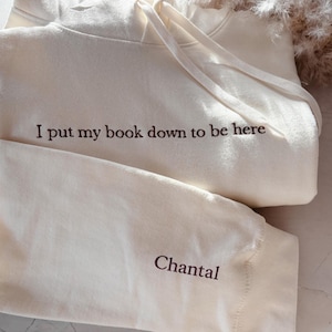 Op de afbeelding: Een lichtgrijs sweatshirt met de tekst "I put my book down to be here." De gevouwen crewneck sweatshirt ligt naast een open boek. De achtergrond is een zachte, neutrale kleur.