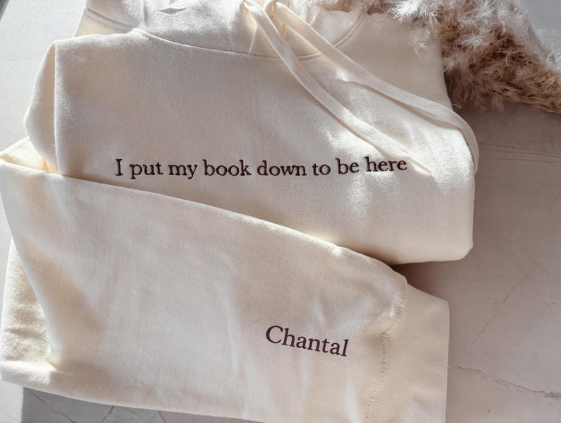 Op de afbeelding: Een lichtgrijs sweatshirt met de tekst "I put my book down to be here." De gevouwen crewneck sweatshirt ligt naast een open boek. De achtergrond is een zachte, neutrale kleur.