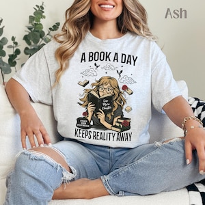 Pode incluir: T-shirt cinza claro com a frase "A BOOK A DAY KEEPS REALITY AWAY". O design apresenta uma pessoa lendo um livro, rodeada por livros, rosas e nuvens. A camisa é cinza claro e tem a palavra "Ash" no canto superior direito.