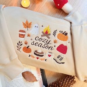 Geborduurde beige sweatshirt met het opschrift 'Cozy Season' en een herfstig borduurontwerp van een kampvuur, pompoenen, spookje, pumpkin spice koffie, warme chocolademelk, herfstbladeren, taart, gebreide muts en winterlaarsjes. De sweater ligt op een open boek met dennenappels, kaarslicht en herfstdecoratie. Perfecte herfsttrui voor Halloween, cozy season en herfstliefhebbers.