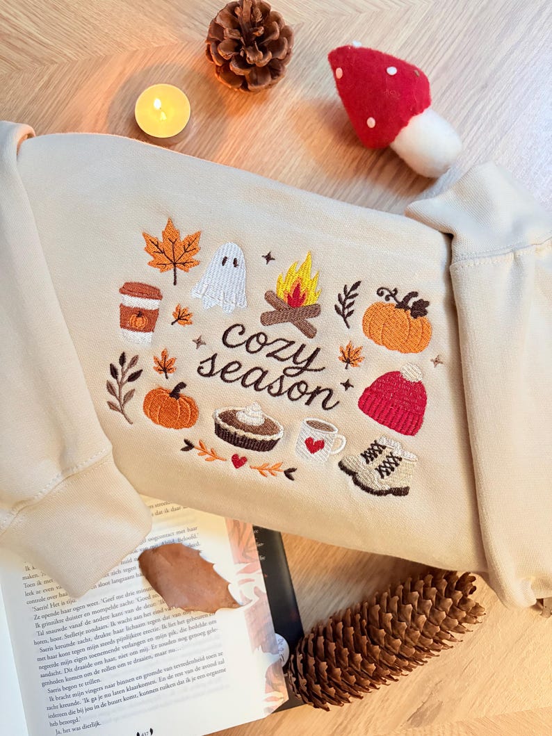 Geborduurde beige sweatshirt met het opschrift 'Cozy Season' en een herfstig borduurontwerp van een kampvuur, pompoenen, spookje, pumpkin spice koffie, warme chocolademelk, herfstbladeren, taart, gebreide muts en winterlaarsjes. De sweater ligt op een open boek met dennenappels, kaarslicht en herfstdecoratie. Perfecte herfsttrui voor Halloween, cozy season en herfstliefhebbers.