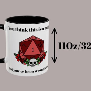 May include: White ceramic mug with a black handle and rim, featuring a red D20 die design with a skull and floral accents. The mug has the text "You think this is a mug... But you've been wrong before." It holds 110 oz (325ml).