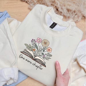 Puede incluir: Sudadera de color crema con un diseño floral que emerge de un libro abierto. El diseño incluye flores rosas, melocotón y amarillas con hojas verdes. El texto "One more chapter" está escrito debajo del libro. La sudadera está doblada sobre una superficie de madera.