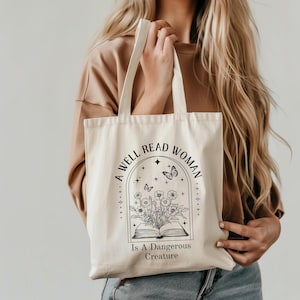 Op de afbeelding: Crèmekleurige draagtas met zwarte tekst en afbeelding. De tekst luidt "A Well Read Woman Is A Dangerous Creature" rond een afbeelding van een open boek met bloemen en vlinders. De tas heeft twee handvatten.