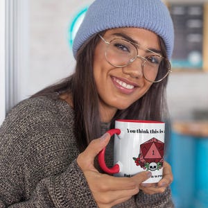 May include: White ceramic mug with a red interior and handle. The mug features the text "You think this is" and a graphic of a red D20 die, flowers, and a skull. The mug is a novelty item.