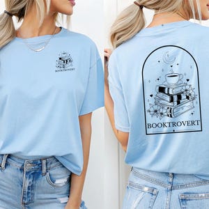 Op de afbeelding: Lichtblauw T-shirt met afbeeldingen voor en achter. De voorkant heeft een stapel boeken met het woord "BOOKTROVERT". De achterkant toont een koffiekopje op boeken, met het woord "BOOKTROVERT" eronder.