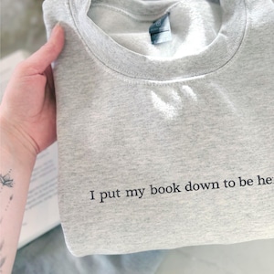 May include: A light gray sweatshirt with the text "I put my book down to be here" printed in black. The sweatshirt is folded and held by a hand. The sweatshirt has a crew neck and a tag.