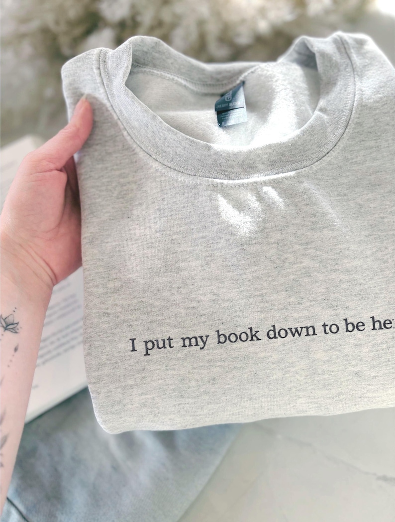 May include: A light gray sweatshirt with the text "I put my book down to be here" printed in black. The sweatshirt is folded and held by a hand. The sweatshirt has a crew neck and a tag.