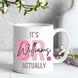 Op de afbeelding: Witte keramische mok met de tekst "It's Dr. Williams Actually" in roze en grijze letters.