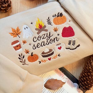 Geborduurde beige sweatshirt met het opschrift 'Cozy Season' en een herfstig borduurontwerp van een kampvuur, pompoenen, spookje, pumpkin spice koffie, warme chocolademelk, herfstbladeren, taart, gebreide muts en winterlaarsjes. De sweater ligt op een open boek met dennenappels, kaarslicht en herfstdecoratie. Perfecte herfsttrui voor Halloween, cozy season en herfstliefhebbers.